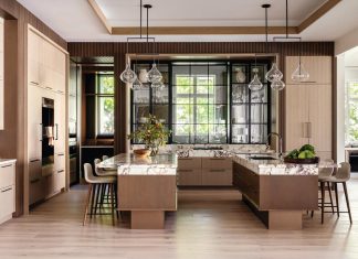 The kitchen’s twin islands appear to float atop walnut-stained cerused oak and plinth supports clad in textured copper. Visual Comfort lighting fixtures illuminate the marble surfaces, which are heavily veined in a deep wine hue. Fluted tambour wood panels trim the perimeter.