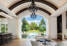 Custom Elegance in Estates at Bay Colony Golf Club Home Bent walnut radius beams sweep across the ceiling, concealing air systems and allowing the architecture by John Cooney to read as clean, continuous, and intentional. Chandeliers are wrapped in black leather, introducing a tactile contrast that softens the scale. Integrated lighting is tucked seamlessly into the structure, eliminating visual clutter and keeping the focus on form and material rather than mechanical elements. The effect draws the eye forward to the dramatic window at the back of the great room, engineered as a single uninterrupted plane of glass.