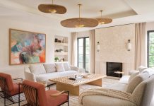 The living room features a coral stone fireplace accented with fine fluting at the lower portion for textural appeal. Built-in shelves flank the artwork centered behind a Poltrona Frau sofa. The abstract painting inspired the subtle orange hues of a pair of Avenue Road lounge chairs and the gold-finished rattan pendants by Hudson Valley Lighting.