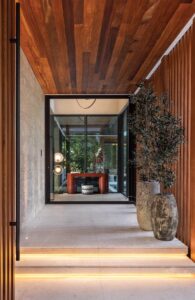A custom glass pivot door framed in warm wood creates a dramatic, almost ceremonial, arrival experience, establishing the home’s contemporary character and refined material palette. Timeless natural textures, including coral stone, natural wood, glass, and custom millwork, create a modern aesthetic that feels both elegant and welcoming.