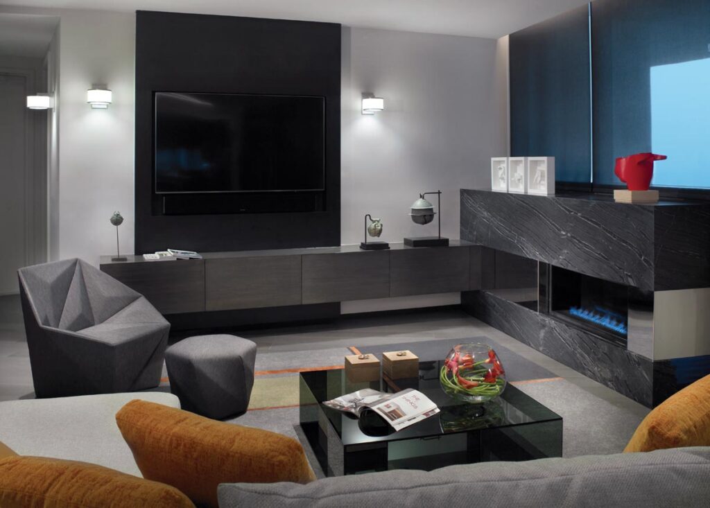 An intimate, darker retreat tailored for family togetherness, this cozy space features a plush Tomlinson Companies sofa and an angled Luminaire chair with a comforting fireplace. A sleek Addison House glass cocktail table maintains a sophisticated edge, while custom cabinetry and a fireplace custom designed by Wolk Design were expertly fabricated by Miami-based Bon Vivant.