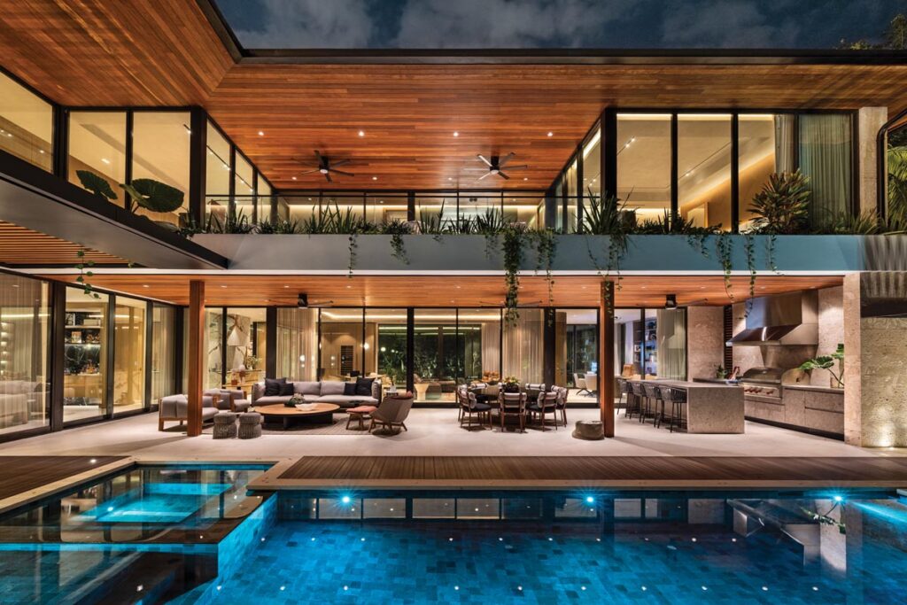 As evening falls, carefully integrated architectural and landscape lighting accentuate the wood ceilings, stone surfaces, and tropical plantings. The home’s pool, conceptualized by CMA Design Studio, shimmers in the foreground.