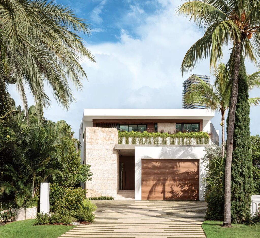 At the front entrance of the residence, a motor court leads to a coral stone entry tower, the narrowest point of the lot, where the architecture is deliberately compressed. The façade presents a monumental coral stone wall flanked by louvers, with a broad step leading up to the entry threshold.