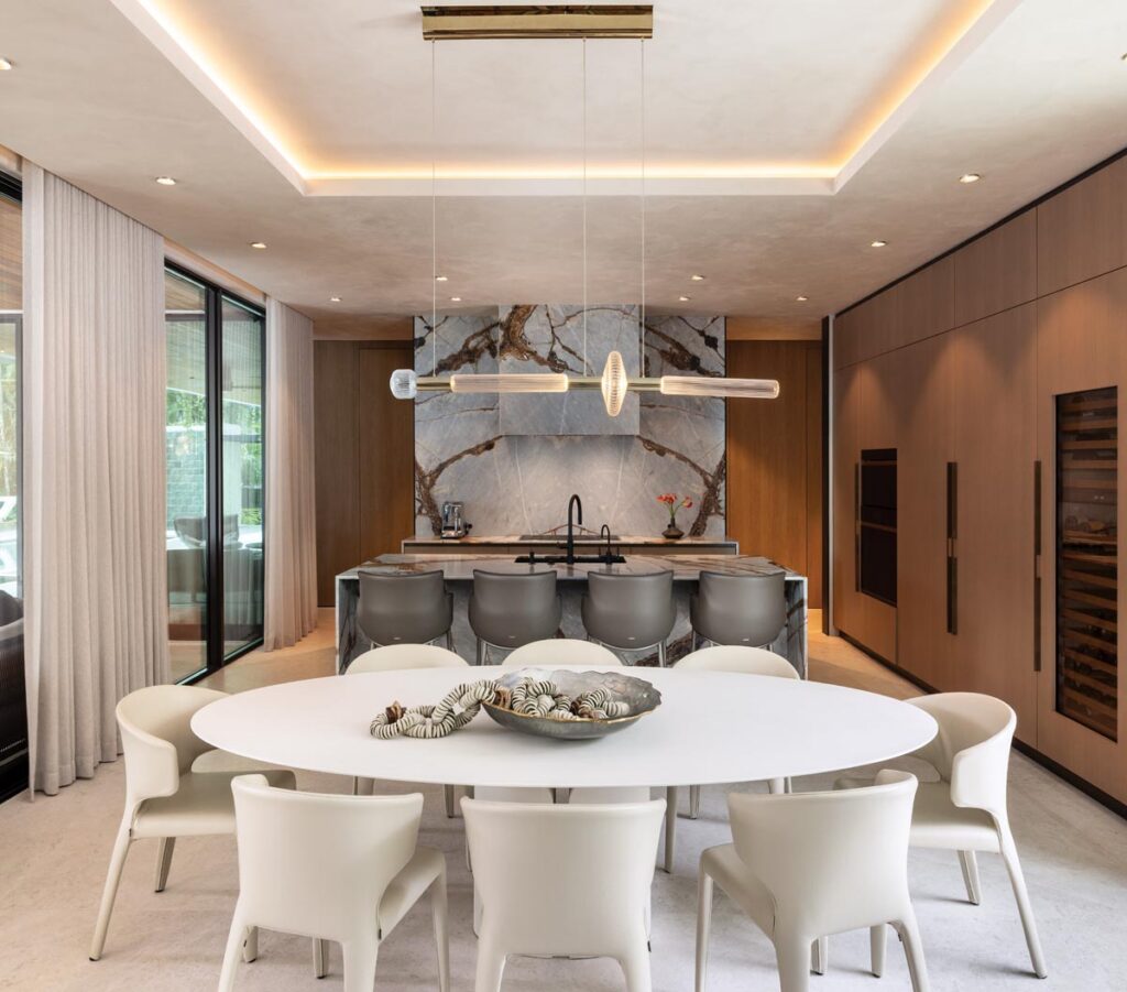 Custom millwork, natural stone surfaces, and fully integrated appliances define the kitchen, creating a highly functional yet elegantly understated space. Fadecci Lighting’s linear chandelier illuminates a waterfall island dotted with Rhianna ML counter stools from Cattelan Italia. MDF Italia’s white NVL dining table, paired with Hola chairs from Cassina, creates a crisp focal point.