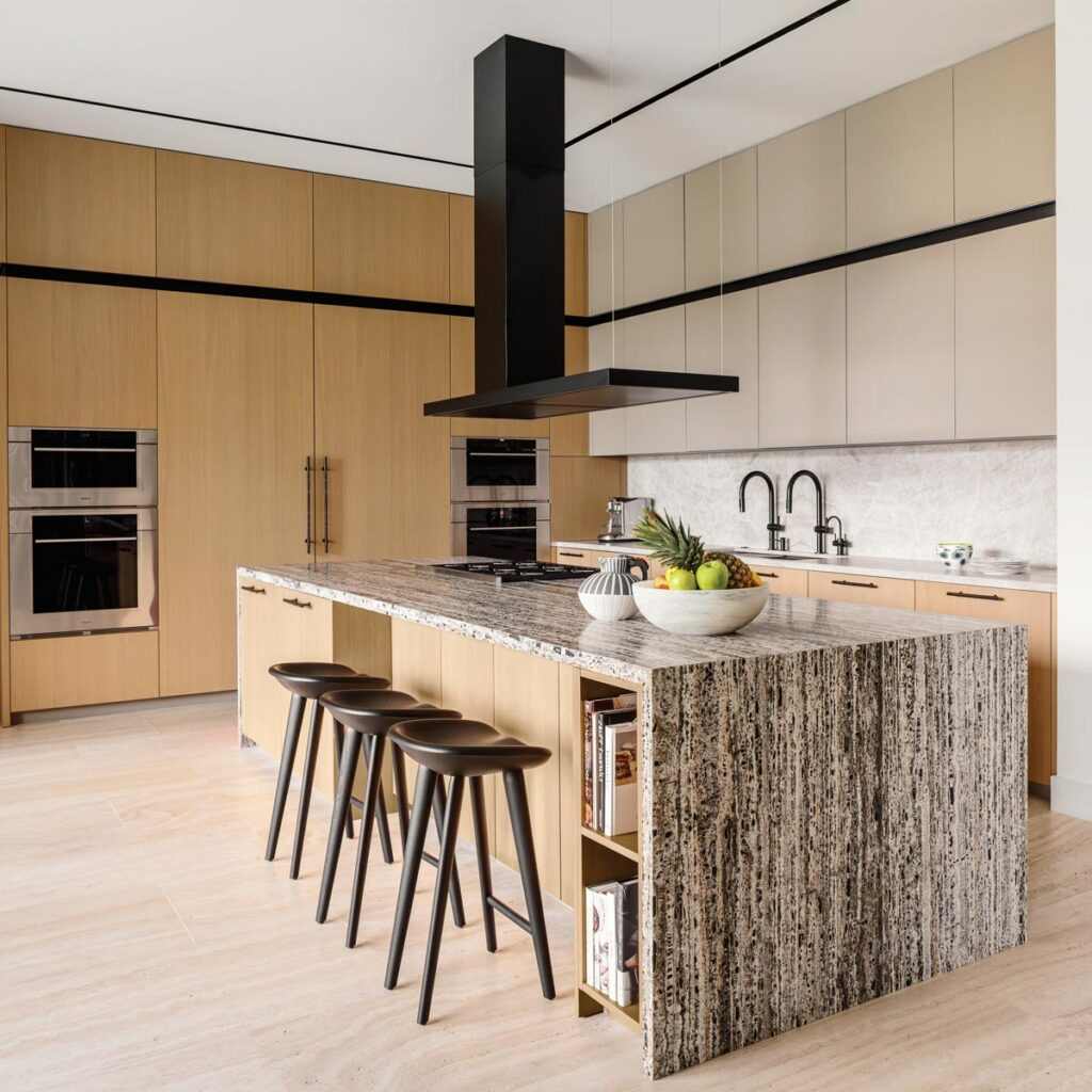The kitchen is the functional and social heart of the home, organized around a generous island clad in black travertine, which serves as the room’s visual centerpiece and provides ample space for gathering and meal preparation. Cabinetry and full-height oak millwork by Interius create a warm, minimalist backdrop, elevated by DWR counter stools and a blackened-steel range hood by Futuro Futuro.