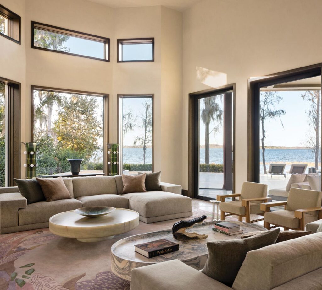 A different perspective of the grand living room showcases the lake view that convinced the homeowners to purchase the property. The taxidermy alligator head nods to the wildlife in the area. Furniture here includes Agatha 01 armchairs by Martin Massé, Derk sofas by Studio Piet Boon, a marble coffee table by Francesco Balzano, and Palm floor lamps by Blue Green Works.