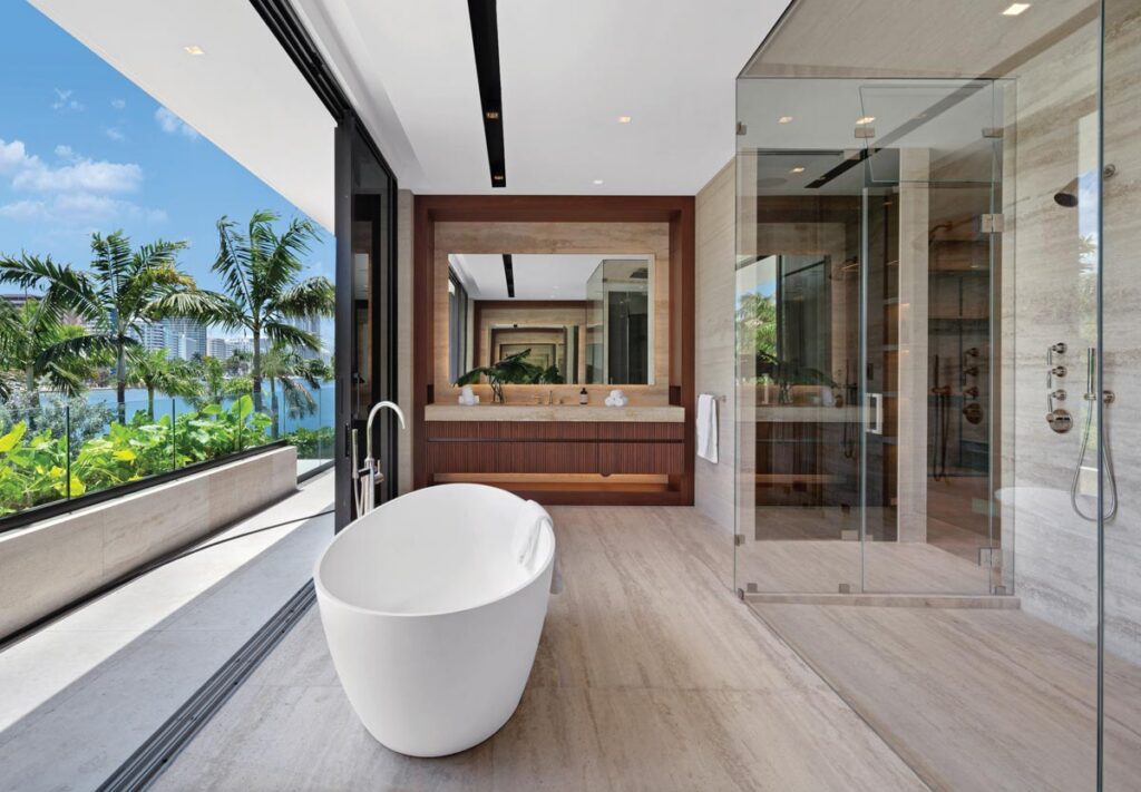 A freestanding soaking tub from Badeloft sits directly before a full-height glazed wall open to the waterway. A deep walnut-framed recess housing a wide backlit mirror, the vanity wall encompasses fluted walnut cabinetry with a travertine countertop. A large walk-in shower in the same travertine is enclosed in frameless glass.