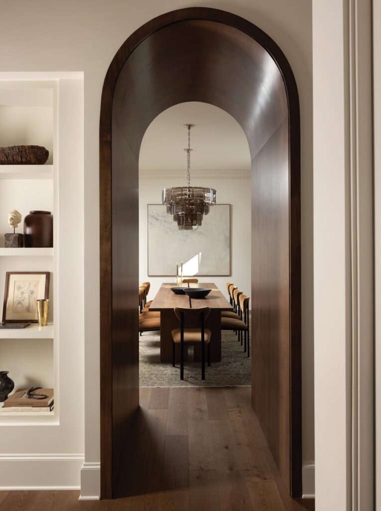 A wood archway frames the dining room while intentionally narrowing the field of view. The room itself is marked by rich tones, sculptural forms, and collected details that give it personality. Funishings includes a table from CFC, chairs from Maiden Home, and a chandelier from Ethan Allen.