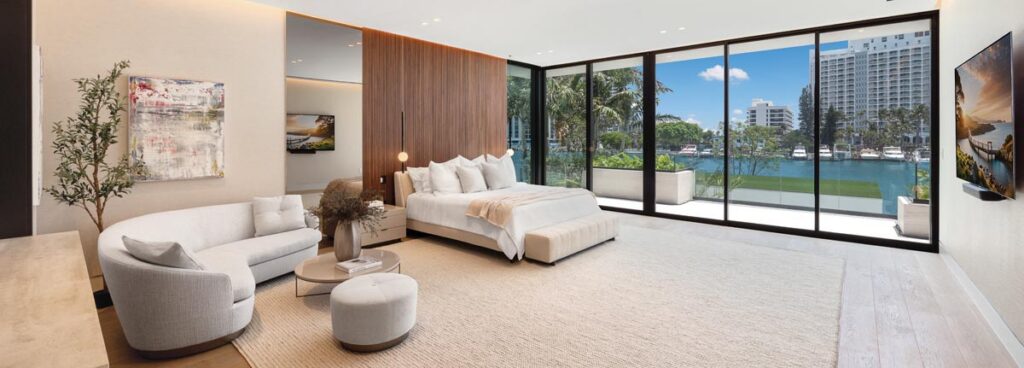 Occupying the full width of the upper level, the primary suite features floor-to-ceiling sliding glass doors that reveal the waterway. Minotti’s upholstered Spencer bed centers on a fluted walnut panel that anchors the room. A sitting area includes Minotti’s Jacques sofa and ottoman.