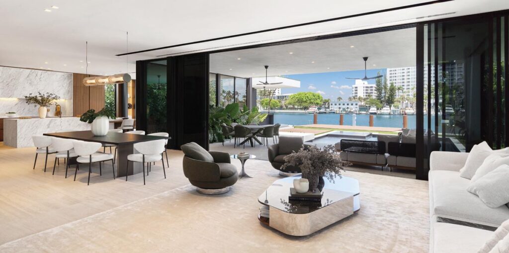 The residence’s ground floor ceiling peaks at over 20 feet, then eases into a more intimate plane as you move toward the water. Floor-to-ceiling black steel sliding panels retract completely, dissolving indoor/outdoor boundaries. Sleek Minotti furnishings include a low-slung ivory sofa, sculptural swivel chairs, and a reflective glass-and-bronze cocktail table.
