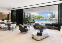 The residence’s ground floor ceiling peaks at over 20 feet, then eases into a more intimate plane as you move toward the water. Floor-to-ceiling black steel sliding panels retract completely, dissolving indoor/outdoor boundaries. Sleek Minotti furnishings include a low-slung ivory sofa, sculptural swivel chairs, and a reflective glass-and-bronze cocktail table.