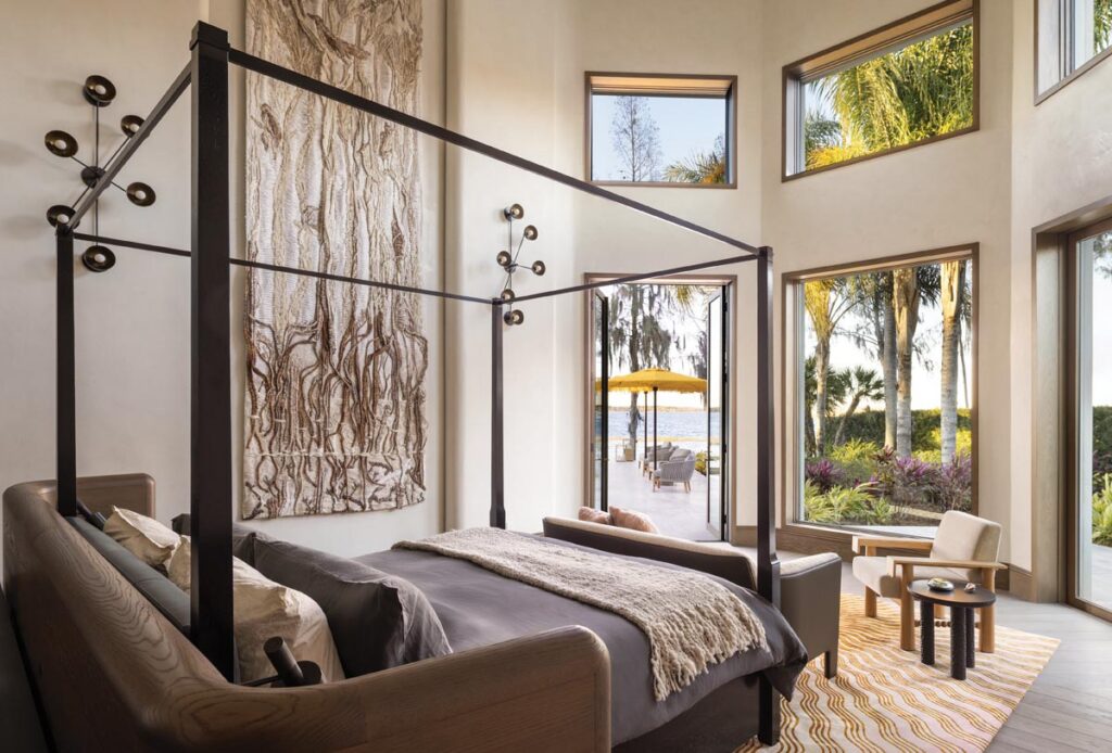 The scale of the primary bedroom was addressed with a poster bed from Holly Hunt and a woven tapestry by KRJST Studio that depicts the marshes of Central Florida. The sconces on the wall are by Apparatus.