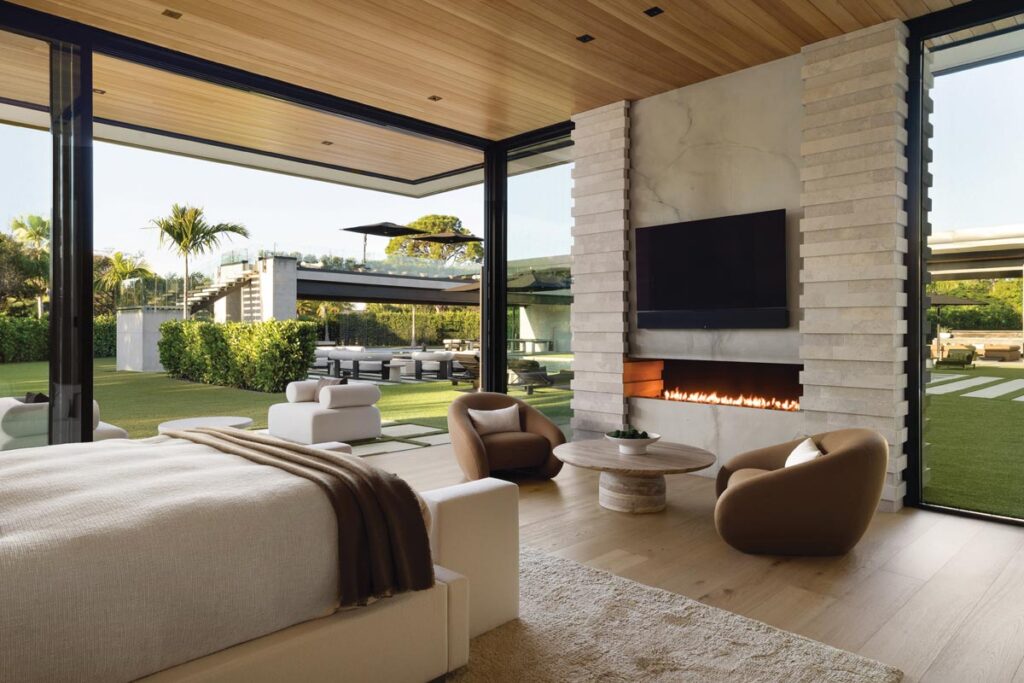 Softness meets structure in the primary bedroom thanks to a monochromatic neutral palette. Cream marble and stone surround the gas fireplace. Curved swivel club chairs and a round cocktail table, all from RH, counterbalance the sharp, linear precision of floor-to-ceiling glass and steel beams.