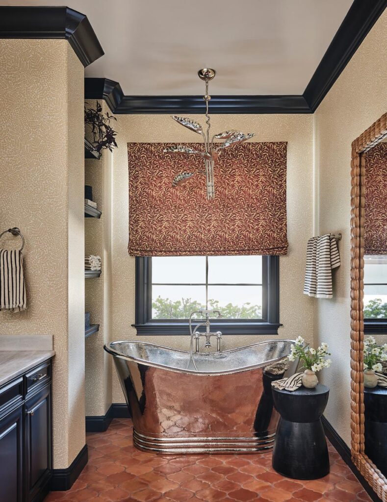 The primary bathroom features a striking satin nickel tub from Vintage Bath & Home complemented by a neutral micro-print wallpaper paired with contrasting dark millwork and cabinetry. A sculptural chandelier from Visual Comfort crowns the space, echoing the tub’s luminous finish.